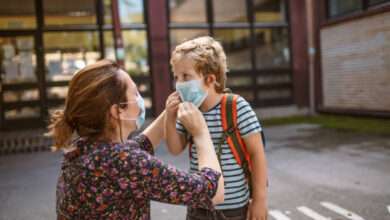 mãe abaixada arrumando a máscara do rosto do filho em frente a escola