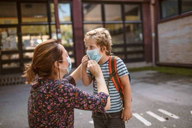 mãe abaixada arrumando a máscara do rosto do filho em frente a escola