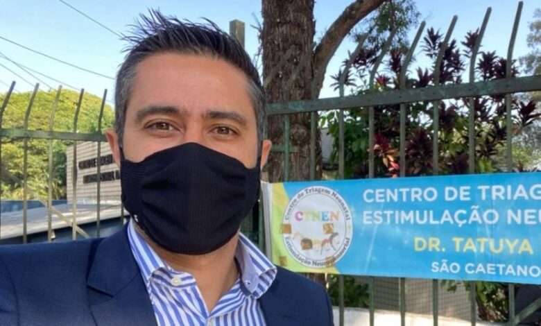 Caio Salgado usando máscara preta e camisa listrada em frente ao CTNEN - Centro de Triagem Neonatal e Estimulação Neurossensorial